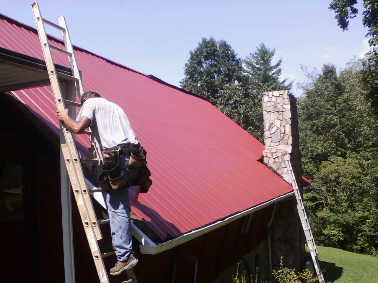 Roofer on a ladder beside a red metal roof and stone chimney — Soffit & Fascia Repair by Roofing Brothers in Saratoga